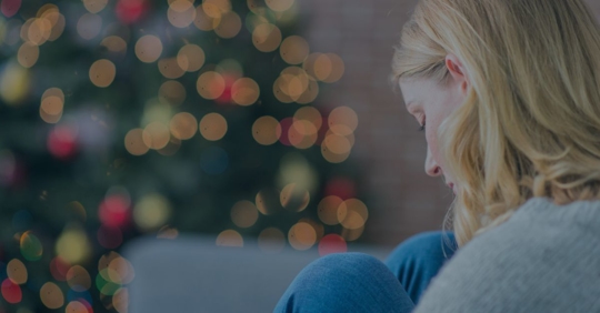 Woman sitting with her knees to her chest and a Christmas tree in the background.